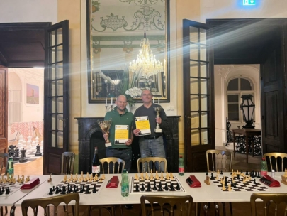 Zwei Männer stehen Seite an Seite in einem eleganten Raum und halten Urkunden und Trophäen in der Hand. Vor ihnen steht ein mit Schachbrettern gefüllter Tisch, und hinter ihnen hängt ein großer Spiegel mit einem Kronleuchter. Die Atmosphäre erinnert an eine Preisverleihung bei einem Schachturnier.