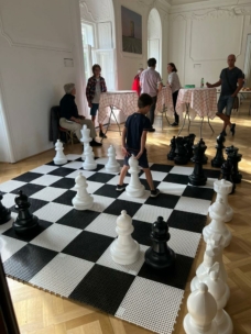 Ein Junge steht auf einem großen Schachbrett mit übergroßen Schachfiguren in einem hellen, eleganten Raum. Im Hintergrund sind mehrere Personen an Stehtischen versammelt, die sich unterhalten und zusehen.