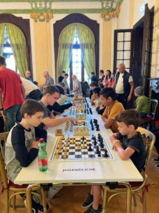 Eine Gruppe von Kindern und Jugendlichen sitzt an Tischen und spielt Schach in einem hell erleuchteten Raum mit großen grünen Vorhängen. Mehrere Erwachsene stehen herum und beobachten die Spiele. Auf dem Schild am Tisch steht JUGENDLICHE.