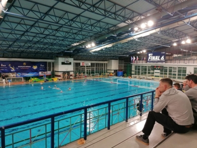 Zuschauer beobachten ein Wasserballspiel in einer Schwimmhalle. Das Becken ist groß, die Spieler sind im Wasser, und im Hintergrund sind Banner mit den Final 6 zu sehen. Die Halle hat ein hohes, blaues Metalldach.