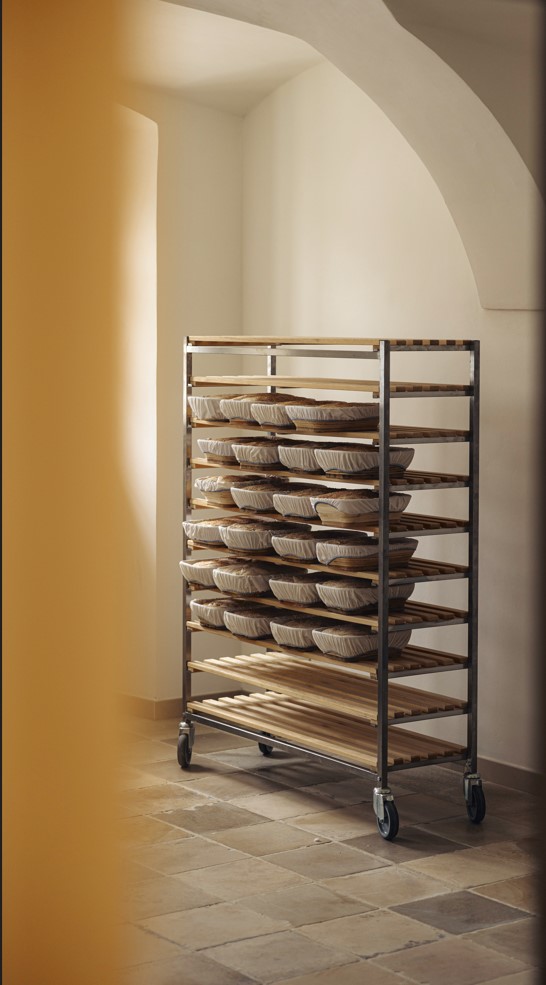 In einem hellen, minimalistischen Raum mit gefliesten Böden und gewölbten Decken steht ein Metallregal mit mehreren Fächern, in denen der Teig in Gärkörben gelagert wird.
