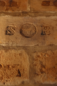 Eine Nahaufnahme eines gealterten Ziegelsteins mit eingravierten Markierungen: S auf der linken Seite, B auf der rechten Seite und 1873 A.D. innerhalb eines Kreises in der Mitte. Die Oberfläche scheint verwittert und strukturiert zu sein.