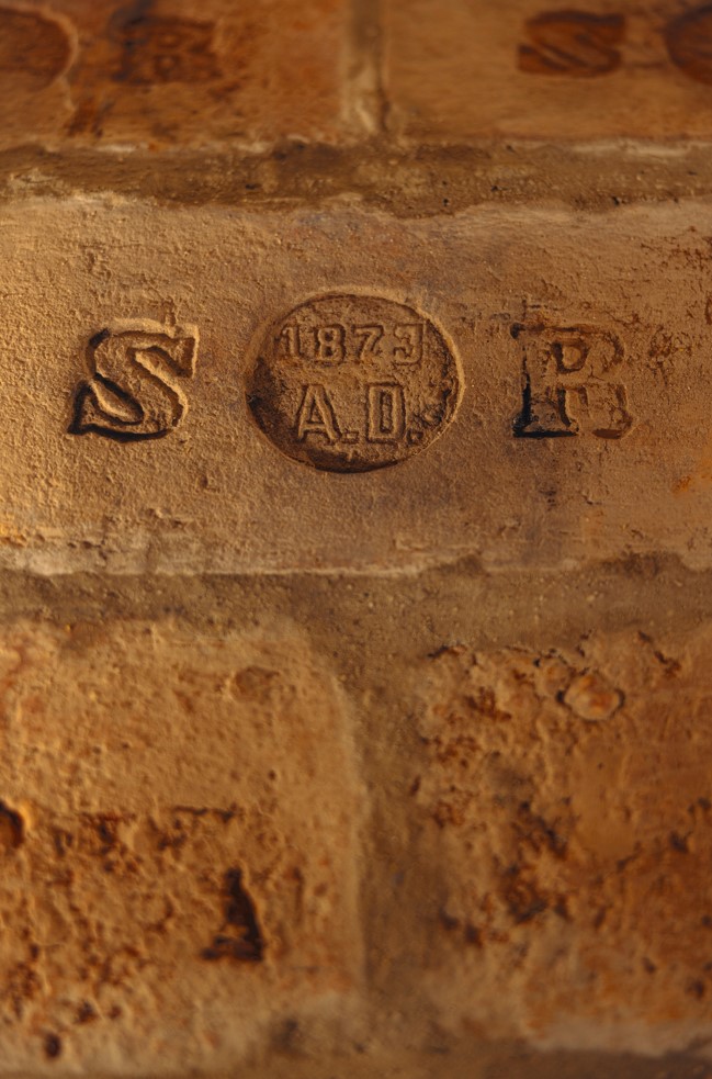 Eine Nahaufnahme eines gealterten Ziegelsteins mit eingravierten Markierungen: S auf der linken Seite, B auf der rechten Seite und 1873 A.D. innerhalb eines Kreises in der Mitte. Die Oberfläche scheint verwittert und strukturiert zu sein.