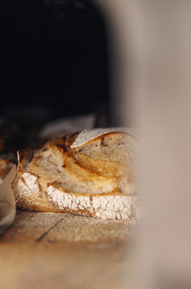 Nahaufnahme eines rustikalen Brotlaibs mit einer rissigen, mehlbestäubten Kruste, der in einem schwach beleuchteten Ofen liegt. Das Brot scheint frisch gebacken zu sein und hat eine goldene, strukturierte Oberfläche.