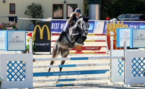 Eine Person mit Helm reitet ein graues Pferd, das bei einem Reitturnier im Freien über ein Hindernis springt. Im Hintergrund sind bunte Absperrungen und Werbung zu sehen.