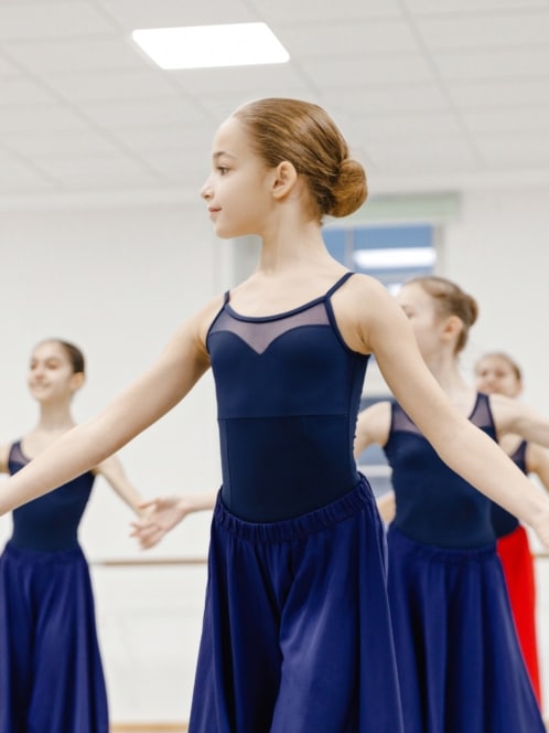 Ein junges Mädchen in einem marineblauen Trikot und Rock steht anmutig in einer Ballettpose, die Arme ausgestreckt, während im Hintergrund in einem hellen Studio andere Mädchen in ähnlichen Outfits tanzen.