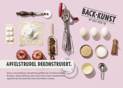 Eine flache Darstellung von dekonstruierten Apfelstrudel-Zutaten, darunter Äpfel, Eier, Rosinen, Zimt, Butter, Zucker, Mehl, Semmelbrösel und ein Schneebesen, auf einem rosa Hintergrund mit deutschem Text, der das Rezept beschreibt.