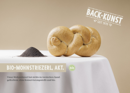 Ein geflochtenes Mohnbrötchen liegt auf einem weißen Tischtuch neben einem kleinen Mohnhaufen. Ein deutscher Text hebt die Bio-Qualität und die handwerkliche Zubereitung des Brotes hervor, darüber ein Back-Kunst seit 1970-Etikett.