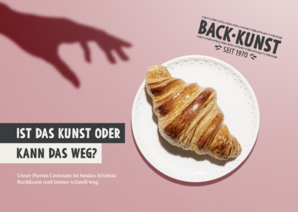 Ein goldenes Croissant liegt auf einem weißen Teller vor einem rosa Hintergrund. Der Schatten einer Hand greift nach dem Croissant. Der deutsche Text fragt, ob es sich um Kunst handelt oder ob es weggenommen werden kann, und verweist auf die Bäckerei Backkunst.
