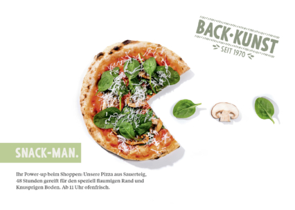 Eine aufgeschnittene Pizza mit Grünzeug, Käse und Tomaten liegt auf einem weißen Hintergrund, der an die Figur Pac-Man erinnert. Spinatblätter und ein Pilz sind daneben angeordnet. Im Bild erscheinen ein deutscher Text und das BACK-KUNST-Logo.