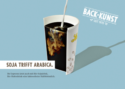 Eine Tasse mit dunklem Kaffee, in die Sojamilch gegossen wird, wodurch ein wirbelndes Muster entsteht. Auf der Tasse sind Kaffeebohnen abgebildet, und der Hintergrund ist blau. Der deutsche Text wirbt für Kaffee mit Soja, Haferdrinks oder laktosefreier Milch.