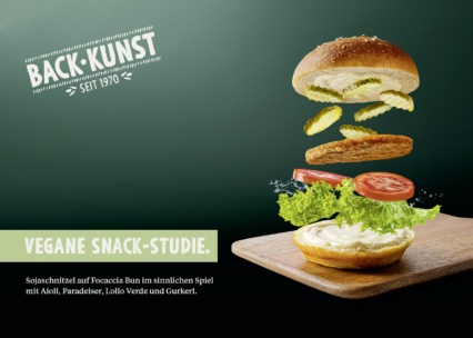 Über einem Holzbrett schwebt ein dekonstruiertes veganes Sandwich mit Focaccia-Brötchenhälften, Salat, Tomate, Essiggurken und einem Aufstrich. Der deutsche Text wirbt für eine vegane Snackstudie von Backkunst.