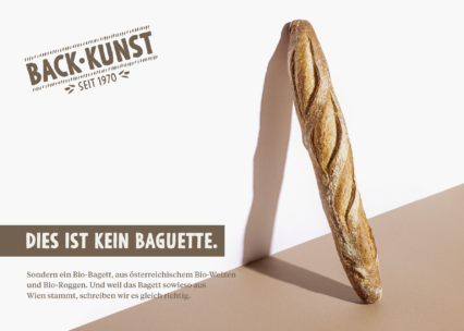Ein einzelnes Baguette liegt diagonal auf einem blassbraunen und beigen Hintergrund. Der Text auf Deutsch lautet: "Dies ist kein Baguette", mit zusätzlichen Angaben zu den österreichischen Bio-Zutaten des Brotes.