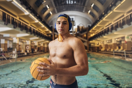 Ein männlicher Wasserballspieler, der eine Badekappe trägt und einen gelben Ball in der Hand hält, steht selbstbewusst am Rande eines Hallenbads, wobei das Becken und die Bahnen im Hintergrund zu sehen sind.