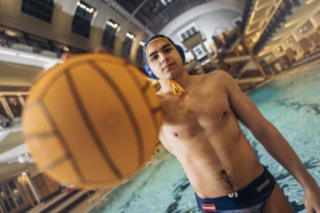 Ein Wasserballspieler mit einer blauen Kappe steht am Beckenrand und hält einen gelben Wasserball in die Kamera, während im Hintergrund ein großes Schwimmbecken und Sprungtürme zu sehen sind.
