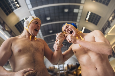 Zwei Männer mit Wasserballmützen stehen in einem Innenraum, beide ohne Hemd, und tun so, als würden sie in dieselbe Eistüte mit Schokoladenüberzug beißen, während sie sie gemeinsam festhalten und spielerische, übertriebene Ausdrücke machen.