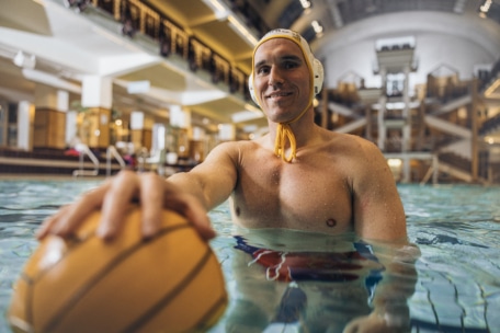 Ein männlicher Wasserballspieler mit Badekappe und Schwimmbrille steht lächelnd in einem Schwimmbecken und hält mit einer Hand einen gelben Wasserball in die Kamera. Die Schwimmhalle ist gut beleuchtet und geräumig.