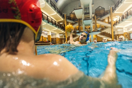 Ein Wasserballspieler mit blauer Kappe bereitet sich darauf vor, den Ball einem Spieler mit roter Kappe zuzuwerfen. Er ist umgeben von Mannschaftskameraden in einem hell erleuchteten Hallenbad mit Treppen und Balkonen im Hintergrund.