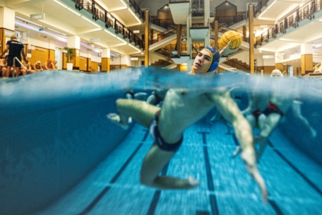Ein Wasserballspieler mit einer blauen Kappe bereitet sich auf den Wurf in einem Hallenbad vor. Das Foto zeigt sowohl über als auch unter der Wasseroberfläche, wobei andere Spieler zu sehen sind, die unter Wasser schwimmen.