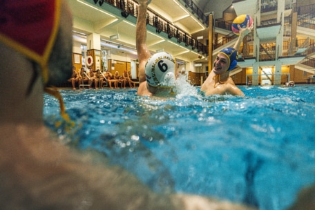 Zwei Wasserballspieler wetteifern in einem Schwimmbecken um den Ball, wobei sich ein Spieler auf den Wurf vorbereitet, während Mannschaftskameraden und Zuschauer von der Seite zusehen. Die Szene ist lebhaft und energiegeladen in einer Schwimmhalle.