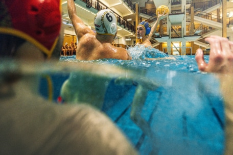 Ein Wasserballspiel in einem Hallenbad zeigt einen Spieler mit blauer Mütze, der den Ball hält und sich auf den Wurf vorbereitet, während ein Gegner mit weißer Mütze den Arm zum Blocken hebt.