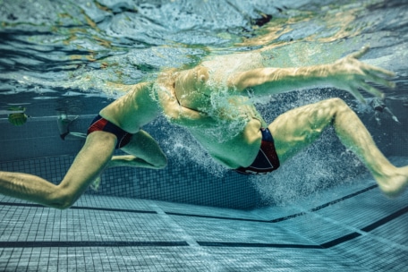 Unterwasseransicht von zwei Schwimmern in Badeanzügen, die nebeneinander in einem Schwimmbecken schwimmen, mit ausgestreckten Armen und Wasser, das um sie herum spritzt. Der geflieste Boden und die Wände des Schwimmbeckens sind im Hintergrund zu sehen.