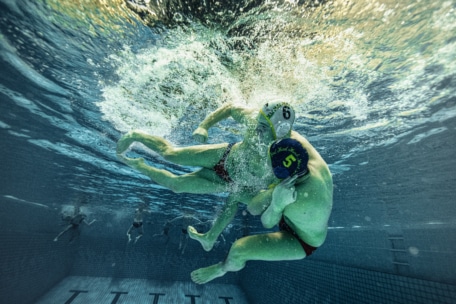 Zwei Wasserballspieler, die Badekappen tragen, kämpfen unter Wasser in einem Schwimmbad und erzeugen dabei Spritzer und Luftblasen, während sie sich während eines Matches heftig bekämpfen. Im Hintergrund sind andere Spieler und Beckenfliesen zu sehen.