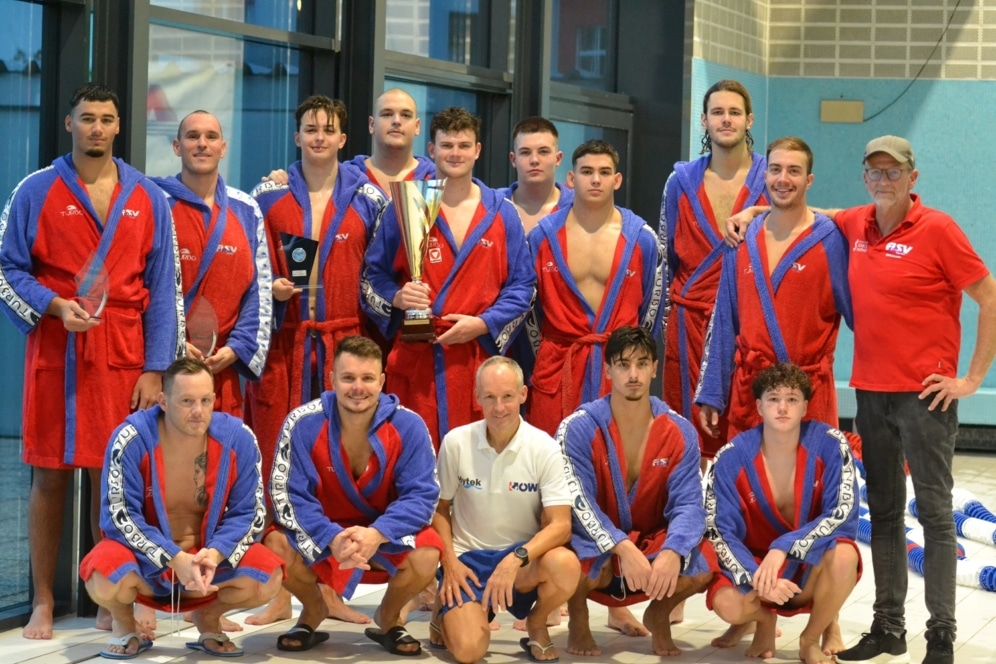 Eine Herren-Wasserballmannschaft posiert in einer Halle und trägt rote und blaue Roben. Zwei Männer im Vordergrund halten einen Pokal und eine Plakette. Zwei Trainer in Freizeitkleidung stehen auf jeder Seite. Alle schauen in die Kamera und lächeln.
