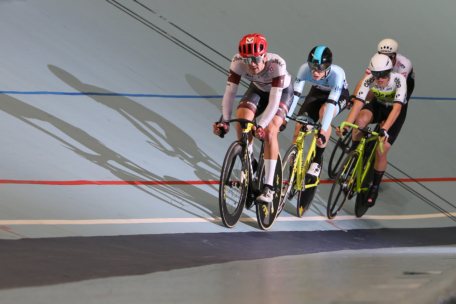 Drei Radsportler in Wettkampfkleidung fahren mit ihren Rennrädern eng beieinander auf einer Indoor-Radrennbahn, lehnen sich in eine Kurve und werfen lange Schatten neben sich.
