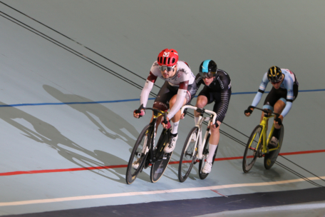 Drei Radfahrer, die Helme und Rennkleidung tragen, fahren auf einer Indoor-Radrennbahn um die Wette. Sie lehnen sich in eine Kurve, während sie eng zusammen fahren. Ihre Schatten sind auf der hellblauen Bahnoberfläche sichtbar.