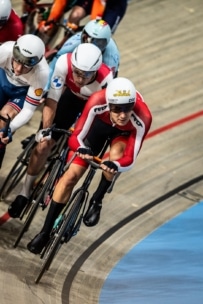 Radfahrer mit Helmen und Rennanzügen treten in einer engen Gruppe auf einer Indoor-Velodrombahn an und lehnen sich während eines Hochgeschwindigkeits-Radrennens in eine Kurve.