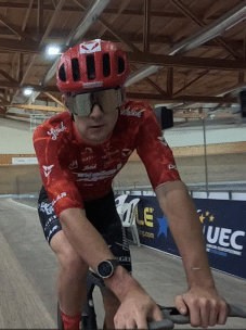 Ein Radfahrer mit rotem Helm und Trikot fährt auf einer Indoor-Velodrombahn. Er trägt eine Sonnenbrille und eine Smartwatch, und im Hintergrund sind Holzbalken und Streckenbegrenzungen mit Schildern zu sehen.