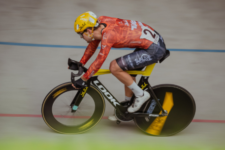 Ein Radfahrer in einem rot-orangenen Trikot und einem gelben Helm fährt mit einem Rennrad auf einer Indoor-Bahn. Der Hintergrund ist unscharf, um die Geschwindigkeit des Radfahrers zu betonen.