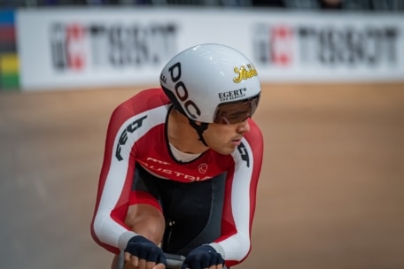 Ein Radfahrer in einem rot-weißen österreichischen Trikot und einem weißen Helm bereitet sich auf ein Rennen auf einer Indoor-Velodrombahn vor. Im Hintergrund ist eine unscharfe Tissot-Werbung zu sehen.