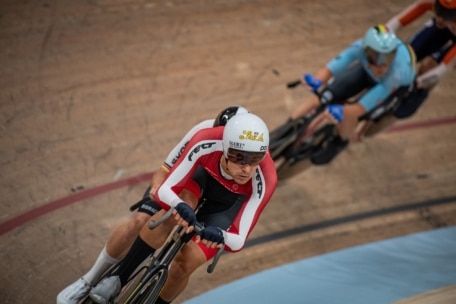 Drei Radfahrer fahren ein Rennen auf einer Indoor-Radrennbahn und neigen sich in eine Kurve. Der führende Radfahrer trägt eine rot-schwarz-weiße Uniform und einen weißen Helm, dicht gefolgt von zwei Radfahrern in blauer und oranger Kleidung.