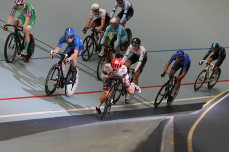 Radfahrer mit Helmen und Rennkleidung fahren auf einem Indoor-Velodrom um die Wette, wobei sie sich in eine schräge Kurve legen. Der führende Radfahrer fährt leicht voraus, während die anderen in einer engen Formation folgen.