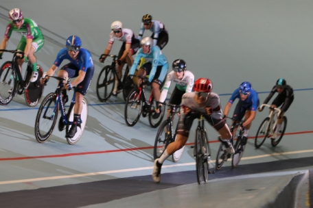 Eine Gruppe von Radrennfahrern in farbenfrohen Uniformen fährt eng beieinander auf einer Indoor-Rennbahn, wobei sie sich in eine Kurve legen, während sie vorwärts fahren.