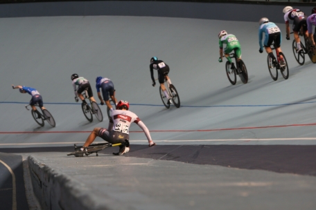 Ein Radfahrer ist gestürzt und rutscht auf der Strecke, während andere Radfahrer auf einem kurvenreichen Velodrom weiterfahren. Der gestürzte Fahrer trägt einen roten Helm und ein weißes Trikot.