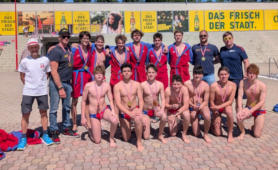 Eine Jungen-Wasserballmannschaft posiert am Pool mit ihren Trainern, die Medaillen und rot-blaue Uniformen tragen. Einige stehen in Bademänteln, andere knien in Badekleidung. Im Hintergrund ist eine gelbe Werbung mit der Aufschrift "DAS FRISCH DER STADT" zu sehen.