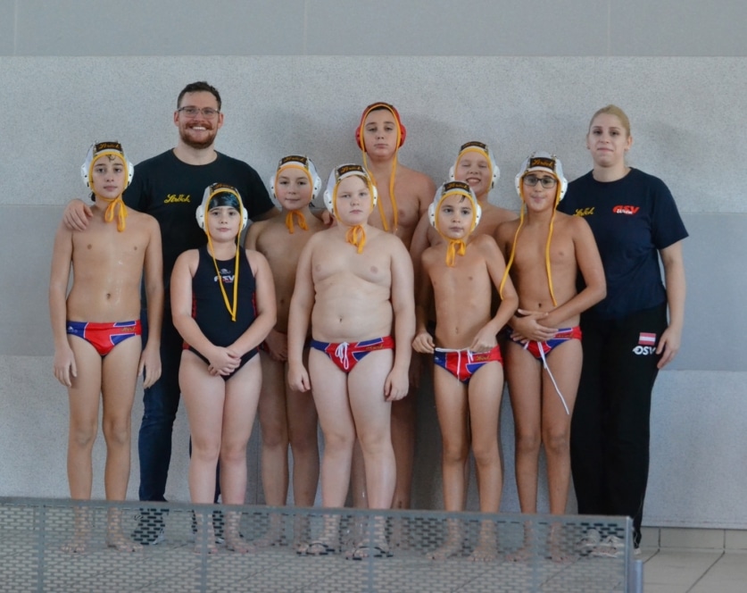 Eine Gruppe von acht Kindern in Badekappen und Badeanzügen posiert zusammen mit zwei erwachsenen Trainern für ein Mannschaftsfoto in einer Halle neben einem Schwimmbecken. Die Mannschaft sieht aus, als wäre sie bereit für ein Wasserballspiel.