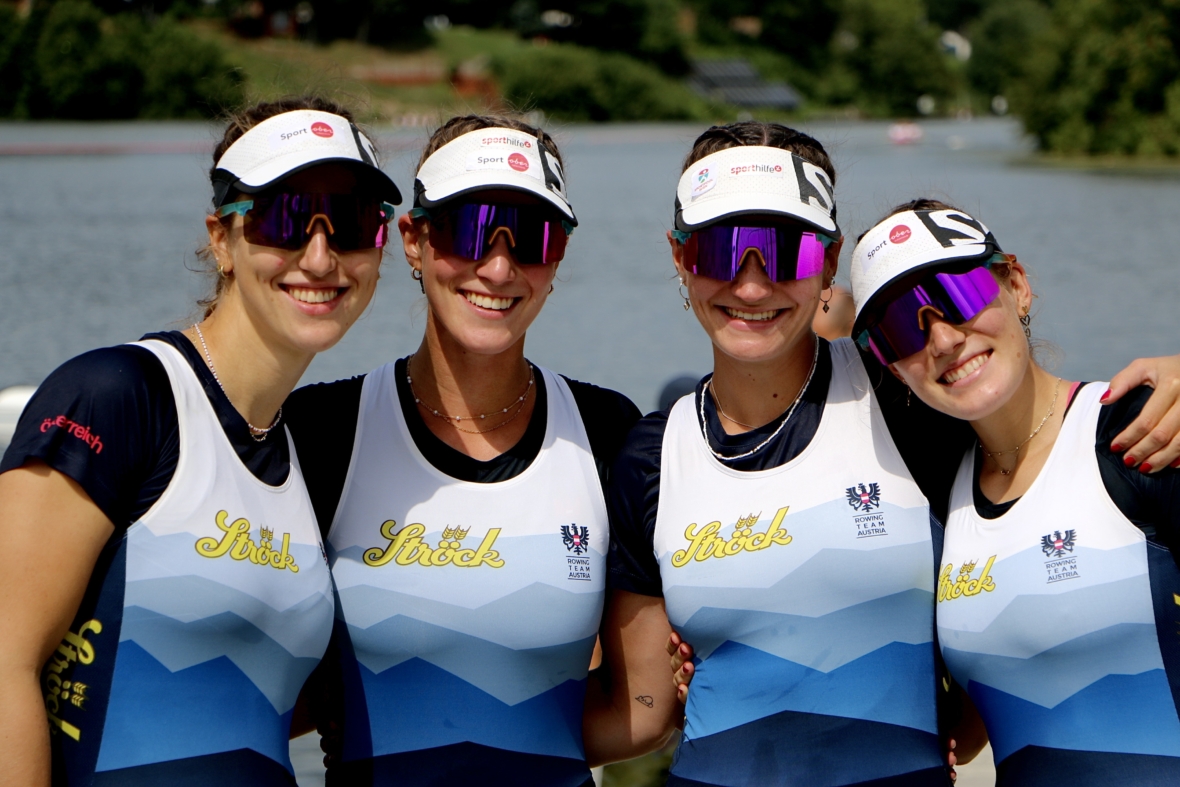 Vier Ruderinnen in passenden Teamuniformen, Sonnenbrillen und Visieren stehen Arm in Arm und lächeln neben einem ruhigen See mit Bäumen und verschwommenen Gebäuden im Hintergrund.