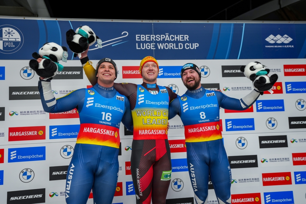 Drei Rennrodel-Athleten in Rennanzügen stehen auf einem Podium, lächeln und halten Panda-Maskottchen, im Hintergrund steht "Eberspächer Rennrodel-Weltcup". Der mittlere Sportler trägt Gelb und Rot, die anderen tragen Blau.