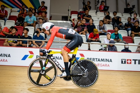 Ein Radfahrer in einem rot-schwarzen Trikot und Helm fährt auf einer Indoor-Velodrombahn, während im Hintergrund Zuschauer von der Tribüne aus zusehen.