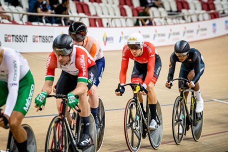 Vier Radsportler fahren ein Rennen auf einer Indoor-Velodrombahn, jeder trägt ein Trikot der Nationalmannschaft und einen Helm. Im Hintergrund sind Reihen leerer roter und weißer Sitze und eine Absperrung mit Sponsorenlogos zu sehen.