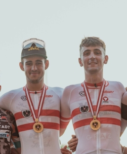 Zwei männliche Radsportler in passenden weiß-roten Uniformen lächeln, während sie nebeneinander stehen, jeder mit einer Goldmedaille um den Hals. Einer trägt eine schwarze Mütze und eine Sonnenbrille auf dem Kopf. Beide haben ihre Arme umeinander gelegt.