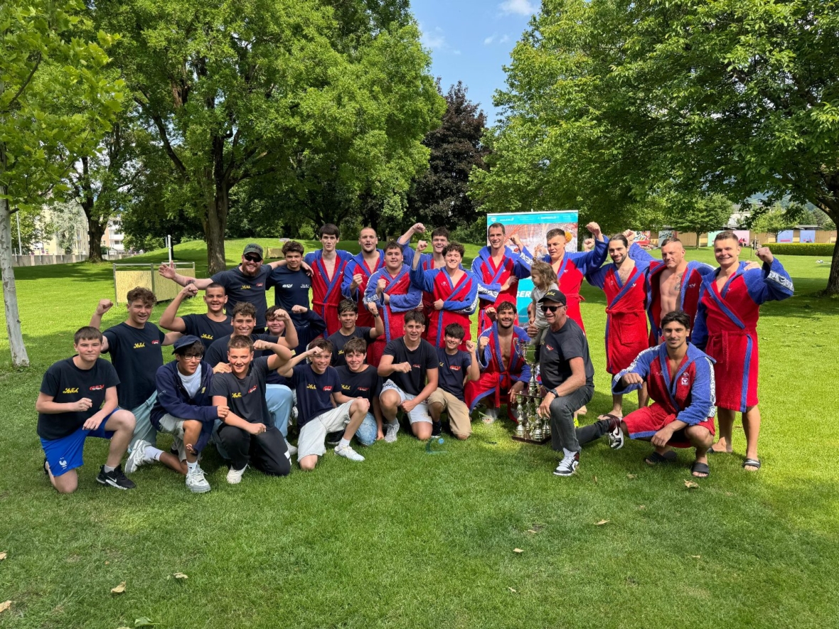 Eine große Gruppe junger Sportler, einige in schwarzen Hemden und Shorts, andere in roten und blauen Sportuniformen, posieren gemeinsam auf einer Wiese mit Bäumen im Hintergrund. Viele bewegen ihre Arme und lächeln.