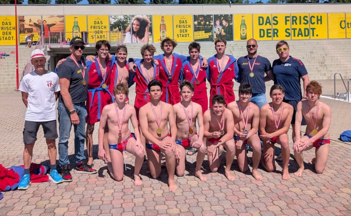 Eine Jungen-Wasserballmannschaft posiert gemeinsam im Freien, mit Medaillen und rot-blauen Uniformen. Einige stehen, andere knien, an den Enden stehen die Trainer. Hinter ihnen hängen gelbe DAS FRISCH DER STADT-Fahnen an einer Wand.