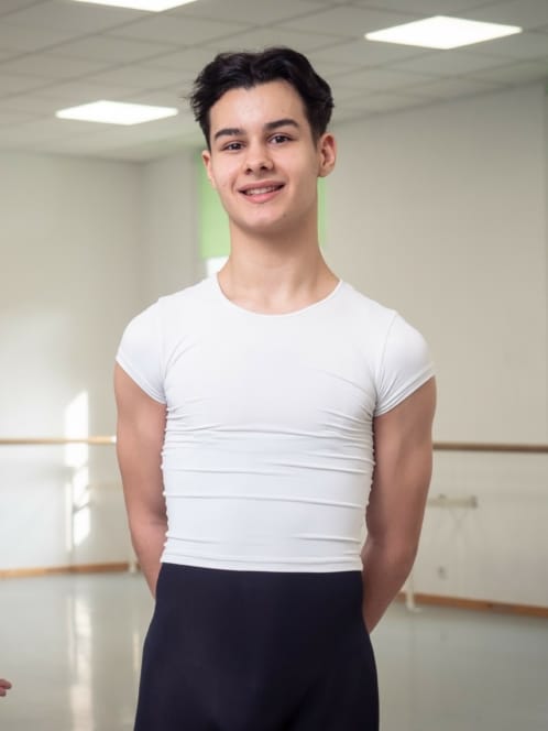 Ein junger männlicher Balletttänzer mit kurzen dunklen Haaren lächelt in die Kamera, während er in einem hellen Tanzstudio steht. Er trägt ein passendes weißes T-Shirt und eine schwarze Tanzhose. Im Hintergrund sind Ballettstangen und große Fenster zu sehen.