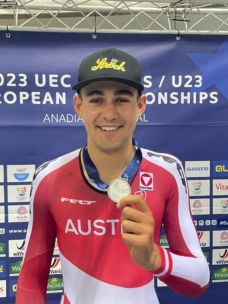 Ein lächelnder männlicher Radsportler im Österreich-Trikot hält eine Silbermedaille der UEC U23-Europameisterschaften 2023 in der Hand, während er vor einem blauen Veranstaltungshintergrund steht. Er trägt eine schwarze Mütze und seine Medaille hängt an einem blauen Band.