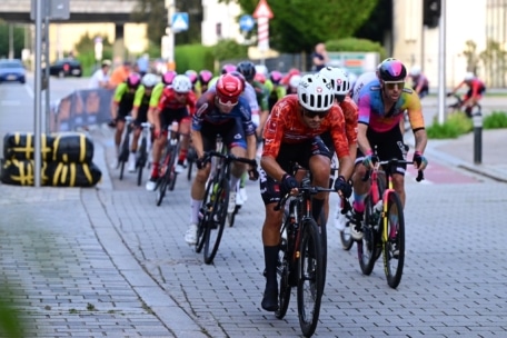 Eine Gruppe von Radfahrern in farbenfrohen Trikots und Helmen fährt auf einer gepflasterten Stadtstraße eng beieinander, wobei sie sich in eine Kurve mit seitlich sichtbaren Absperrungen legen.